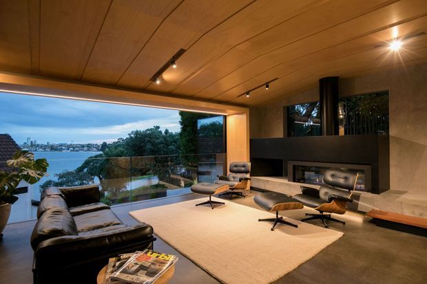 A sophisticated living room with a warm timber-lined ceiling, full-height corner glazing, and expansive waterfront views. The room features a dark leather sofa, mid-century lounge chairs with ottomans, a large pale area rug, a sculptural black fireplace surround, and a long timber bench. Integrated track and recessed lighting create a warm, refined atmosphere.