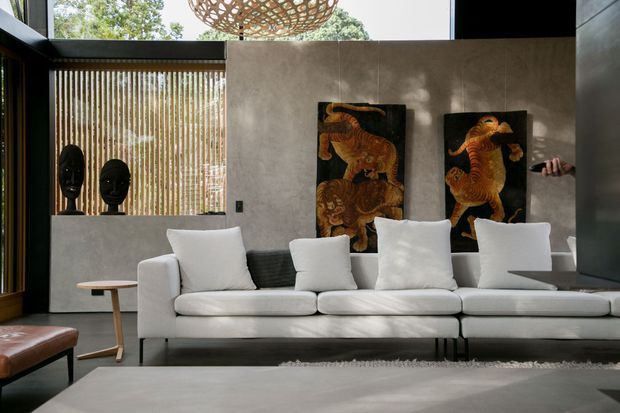 A contemporary living room with a large white sectional sofa facing the camera. The space features dark-framed floor-to-ceiling glazing, vertical timber screening, a grey textured feature wall, black sculptural objects, and two colourful animal artworks. A woven pendant light hangs above, while a low grey coffee table and leather seating complete the refined, dramatic interior.