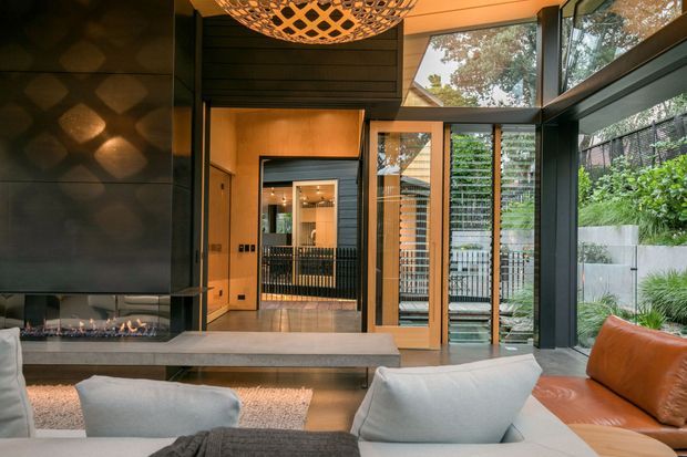 A warm contemporary living room with a dramatic dark feature wall and linear fireplace, facing a glazed entry or hallway and landscaped exterior. Timber-lined walls and ceiling, black-framed floor-to-ceiling glazing, a sculptural pendant light, and pale upholstered seating create a refined indoor-outdoor atmosphere.