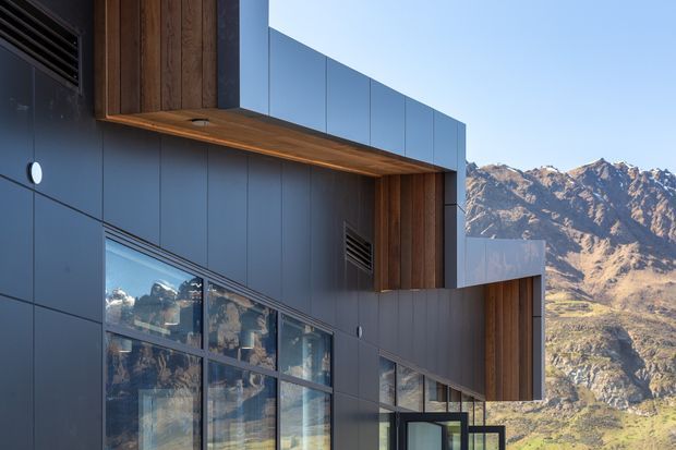Close exterior view of a contemporary multi-level building with dark blue-grey metal cladding, warm vertical timber-lined recesses, projecting box forms, and extensive reflective glazing. Rugged mountains and a sunlit valley are visible beyond and reflected in the windows.