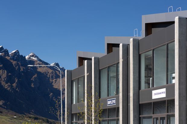 Exterior view of a modern multi-storey commercial building with a stepped roofline, large floor-to-ceiling glazing, dark metal panels, and exposed concrete vertical elements. Business signage is visible on the facade, with a dramatic alpine mountain backdrop.