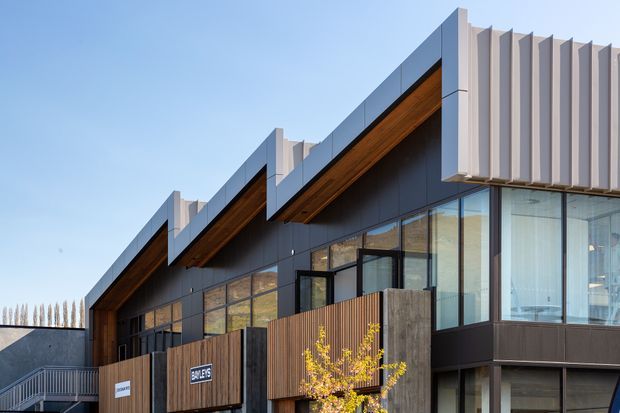 Exterior view of a contemporary multi-level building with dark metal cladding, extensive glazing, warm timber soffits, projecting roof forms, and a prominent vertical fin screen on the upper facade. Timber-clad privacy fencing and an external stair are visible in the foreground.