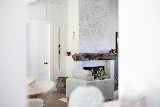 Bright, refined living room with white paneled walls, a white paneled door, a textured light stone or plaster fireplace surround, and a substantial rustic timber mantel. A pale upholstered armchair and sofa face the fireplace, complemented by woven baskets, neutral furnishings, and understated natural decor.