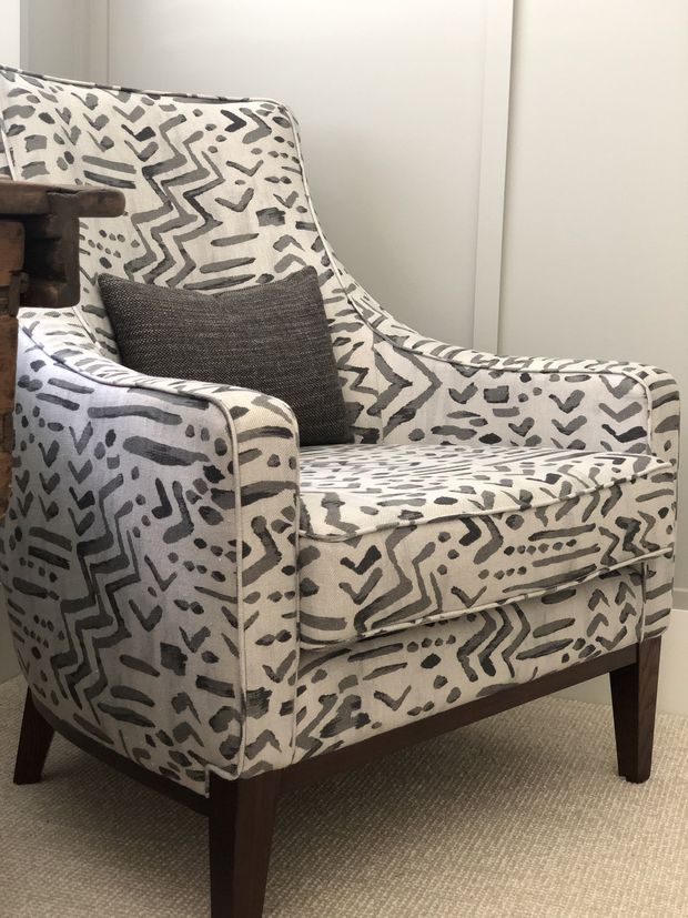 Close-up interior photograph of a traditional upholstered armchair with rolled arms, dark timber legs, and a cream fabric featuring an abstract charcoal-grey geometric pattern. A dark textured cushion rests on the seat, with a neutral wall and carpeted floor in the background.