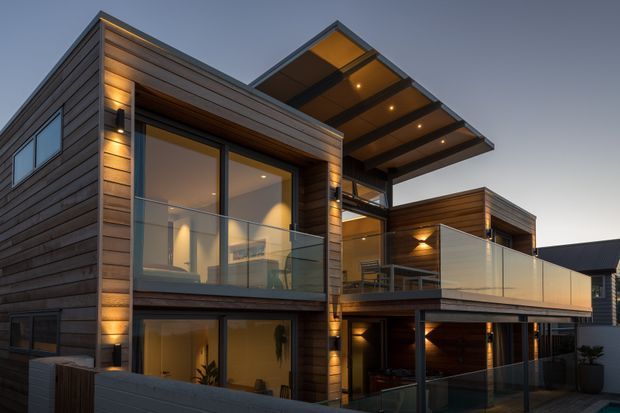 Twilight exterior view of a contemporary multi-level residence with extensive horizontal timber cladding, large glazed openings, recessed warm lighting, cantilevered roof forms, and glass-balustraded balconies and terraces.