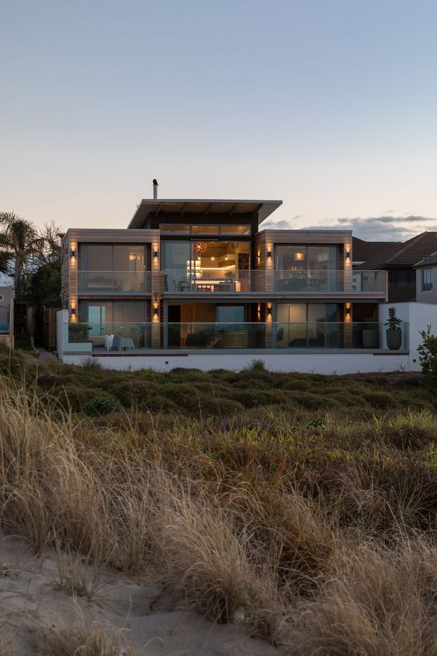 Exterior photograph of a contemporary multi-level coastal residence at dusk. The home features extensive floor-to-ceiling glazing, glass balcony balustrades, warm interior lighting, a projecting flat roof over the upper level, and a timber-toned facade. It is set behind natural dune grasses, with neighbouring houses visible on either side.