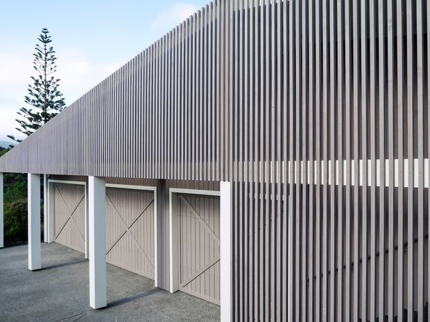 Exterior view of a contemporary building with a long, sharply sloping roofline and extensive vertical slatted screening. Three pale timber-style garage or storage doors sit beneath the overhang, supported by white rectangular posts, with a grey paved driveway in front.