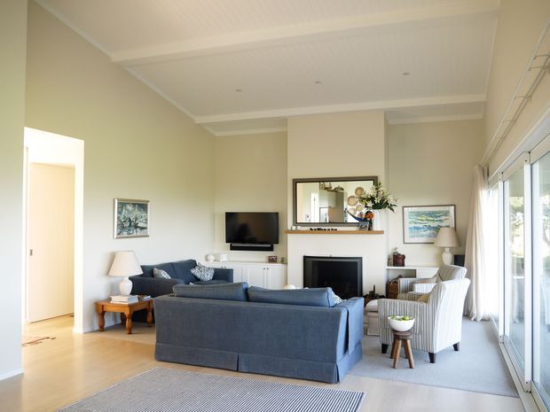 Bright, refined living room with a high coffered white ceiling, pale neutral walls, timber flooring, and expansive glazed doors along one side. A dark blue sofa and matching armchairs face a fireplace and wall-mounted television, with a console mirror, artwork, and restrained decorative furnishings completing the room.