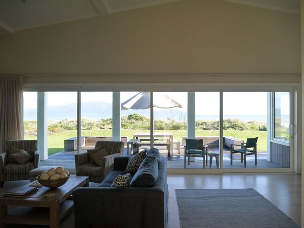 A bright, spacious living room with large floor-to-ceiling sliding glass doors opening onto a furnished outdoor deck and lawn. The room contains a sectional sofa, armchairs, a timber coffee table, and a large rug. Beyond the glazing are a dining table and outdoor chairs, with a coastal landscape and water visible in the distance.