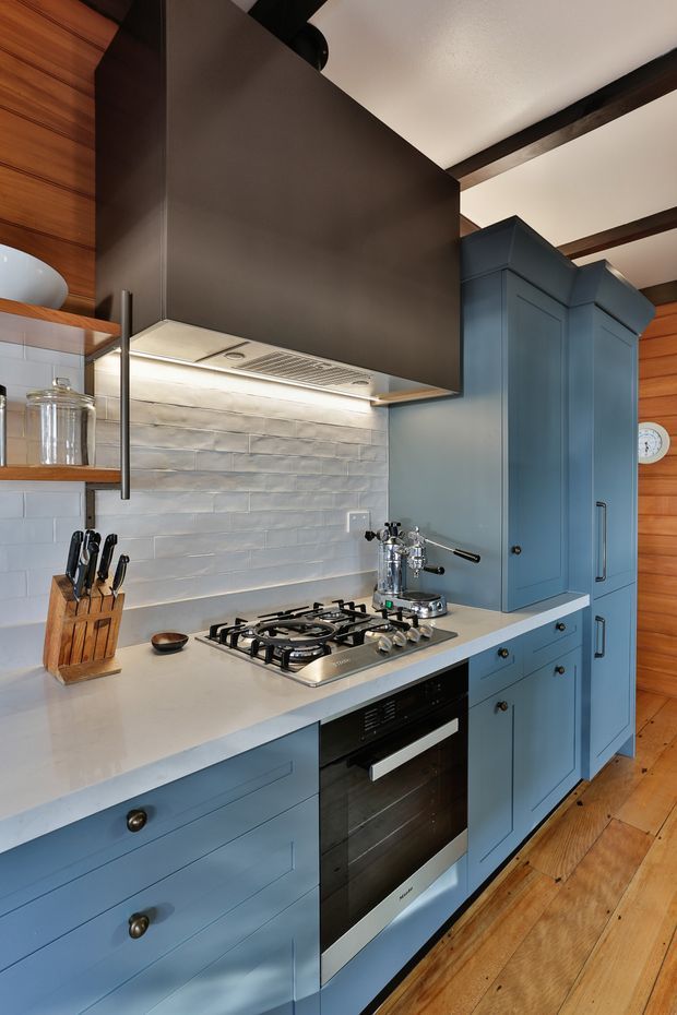 A compact, richly detailed kitchen featuring blue shaker-style cabinetry, a pale engineered-stone benchtop, stainless-steel gas cooktop and oven, and a light grey textured tile splashback. Warm timber wall panelling and timber flooring contrast with the dark metal rangehood canopy and exposed dark ceiling beams. Fixed shelving, a knife block, espresso machine, and other benchtop items reinforce the functional kitchen setting.