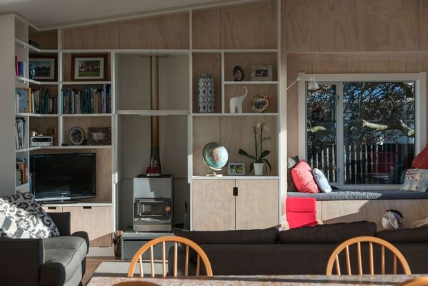 A warm, built-in living room featuring full-height pale timber wall cabinetry and open shelving filled with books, framed art, ceramics and display objects. The joinery incorporates a television, fireplace, storage, and a window-side upholstered bench seat with cushions. Dark sofas and timber dining chairs occupy the foreground, with natural light entering through the window.