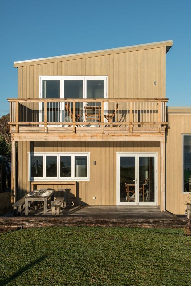 Exterior view of a two-storey timber-clad residential building with a distinctive sloping roofline. The facade features white-framed windows and glazed doors, a substantial upper-level balcony with timber balustrades, and a lower timber deck with outdoor dining furniture. The structure is set against a clear blue sky and lawn.