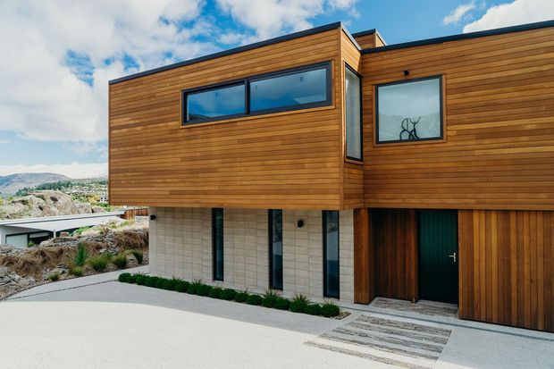 Exterior view of a contemporary multi-level house with extensive horizontal timber cladding, dark-framed glazing, pale masonry lower walls, and a recessed entry beside a timber garage door. The home sits on a sloped, landscaped site with mountain and suburban scenery visible in the background.