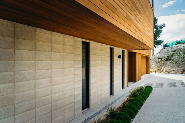 Exterior architectural view of a contemporary home with a long projecting upper volume clad in warm horizontal timber, a recessed timber soffit, and a lower facade of pale concrete blockwork. Narrow vertical dark-framed openings and a timber garage or entry door line a light gravel-paved approach, with minimalist planted edging.