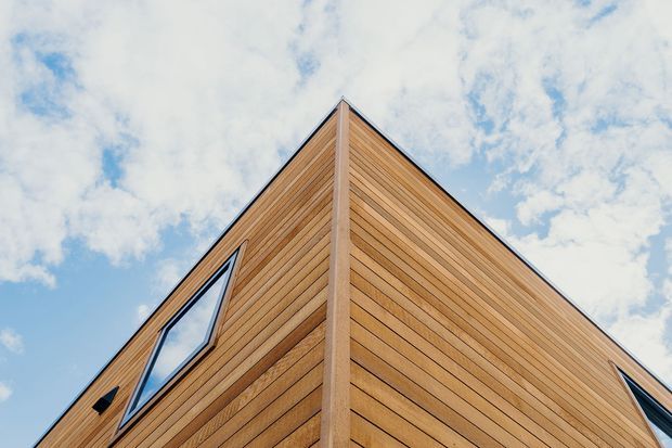 Low-angle exterior view of a sharply peaked, angular building corner clad in warm horizontal timber boards. Narrow dark-framed windows are visible on both facades, with a bright partly cloudy sky behind the roofline. The image emphasizes the geometric gable-like form and timber facade.