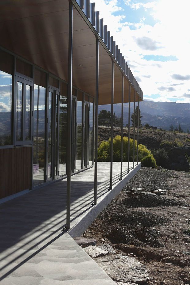 Exterior view of a contemporary, elongated building set on a rugged mountainous site. A covered veranda or walkway runs along the glazed facade, supported by slender dark metal posts and topped with a projecting roof featuring regularly spaced vertical fins. Full-height glazing and glass doors reflect the surrounding landscape. The building has a warm wood-toned soffit and lower wall cladding, with a concrete paving strip and exposed rocky terrain beyond.