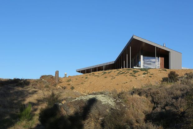 A modern, low-profile house set on a sparsely vegetated, rocky sloping site. The building features a strongly cantilevered roof and projecting upper volume, dark vertical cladding, extensive floor-to-ceiling glazing, and a long covered terrace or wing. Clear blue sky and rugged scrubland surround the isolated residence.