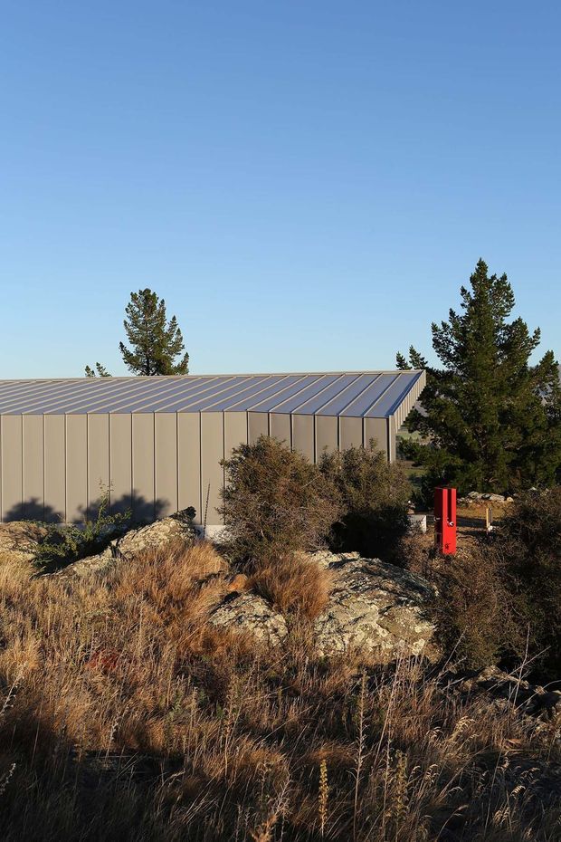 Exterior view of a low, elongated contemporary building set within a rugged, dry natural landscape. The building has a muted beige vertical-panel facade and a shallow sloping standing-seam metal roof, partially screened by shrubs and tall coniferous trees. Rocky terrain and grasses dominate the foreground, with a small bright red vertical exterior element providing contrast.