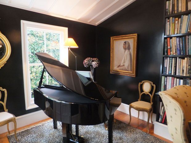 A richly styled traditional music room or living space featuring a glossy black grand piano, dark charcoal walls, white coffered ceiling detailing, built-in floor-to-ceiling bookshelves, a framed portrait, gold-framed upholstered chairs, and a patterned area rug. Daylight enters through a white-trimmed window, creating a refined, dramatic atmosphere.