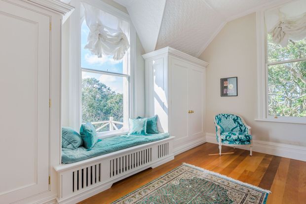 Bright, elegant sitting room with a built-in window seat beneath tall sash windows, a large white paneled wardrobe or cabinet, and a patterned upholstered armchair. The room features polished honey-toned timber flooring, white decorative trim and cabinetry, turquoise cushions and upholstery, and a traditional patterned rug. Abundant daylight and views of leafy trees create an airy, refined atmosphere.