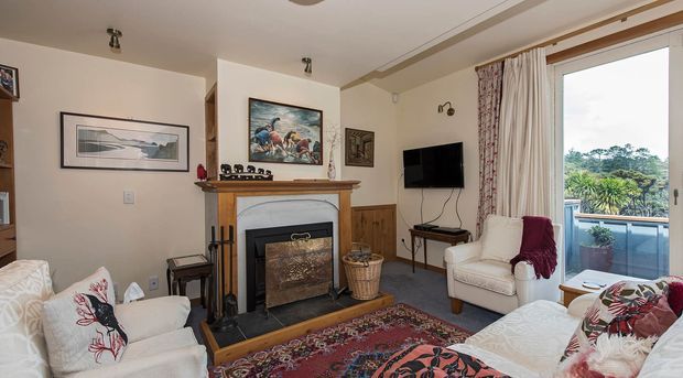 A warm traditional living room with white upholstered seating arranged around a fireplace with a timber mantel and dark firebox surround. The room features wood wall panelling, framed artwork, a patterned Persian-style rug, floral textiles, curtains, and large glazed balcony doors overlooking greenery.