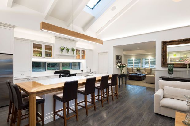 Bright open-plan kitchen and living area with white shaker-style cabinetry, a long timber island with dark bar stools, dark timber-look flooring, exposed white ceiling beams, a large timber ceiling feature, skylights, and a connected lounge with a sofa and framed mirror.