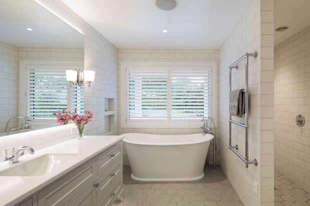 Bright, spacious traditional bathroom with a freestanding white bathtub beneath plantation-shuttered windows, a double vanity with pale grey cabinetry and white countertop, and a tiled walk-in shower. White subway wall tiles, hexagonal floor tiles, chrome fixtures, and a large mirror create a clean, refined look.