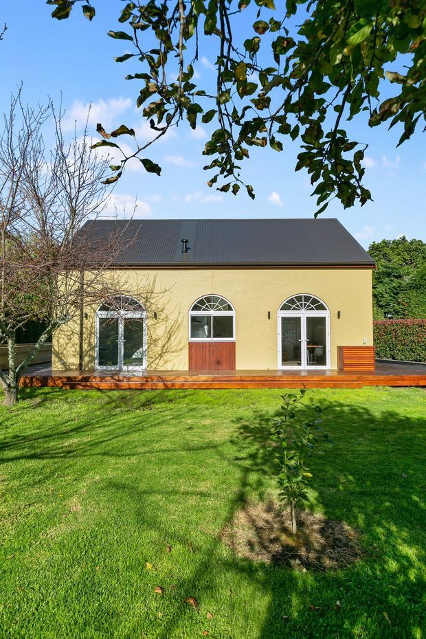 A single-storey yellow rendered house viewed from a landscaped garden. The facade has a dark pitched metal roof, three tall arched-top glazed doors or windows with white frames, and a timber deck with built-in timber seating. Mature trees and lawn surround the building.