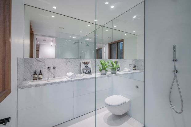 Contemporary bathroom with a wall-hung toilet, frameless glass shower enclosure, large mirrored wall, white glossy built-in vanity cabinetry, and a marble-look wall ledge and backsplash. Recessed ceiling lights and timber-framed openings add warmth to the bright, refined space.