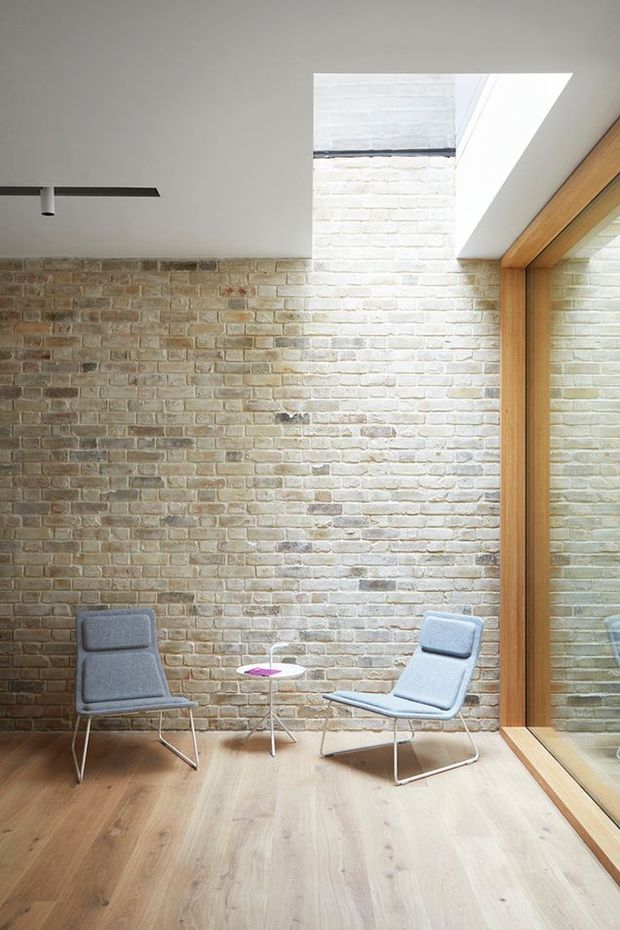 A bright, minimalist sitting area with two grey lounge chairs and a small side table positioned against an exposed light brick wall. The room features a pale timber floor, a large timber-framed glazed opening or mirror on the right, and a roof skylight bringing in strong natural light. A slim black linear ceiling light adds a modern detail.