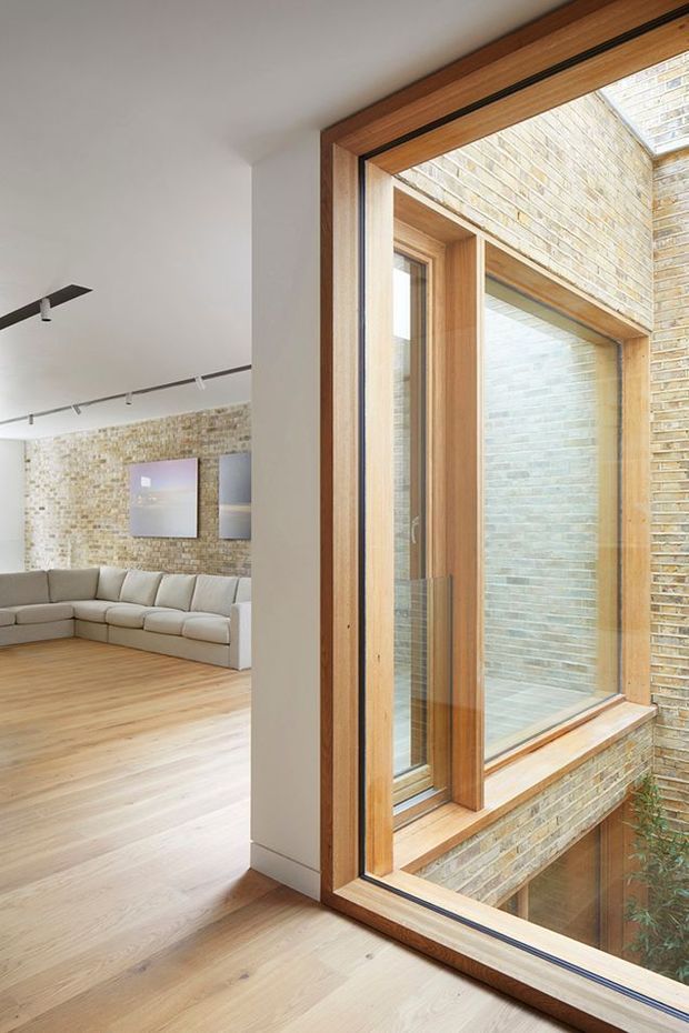 Bright contemporary living room viewed beside a large internal or external glazed opening. The space features pale timber flooring, white walls, a light upholstered sectional sofa, track lighting, and a textured exposed brick feature wall. The prominent glazing has warm timber frames and overlooks a brick-lined lightwell or courtyard, bringing abundant natural light into the room.