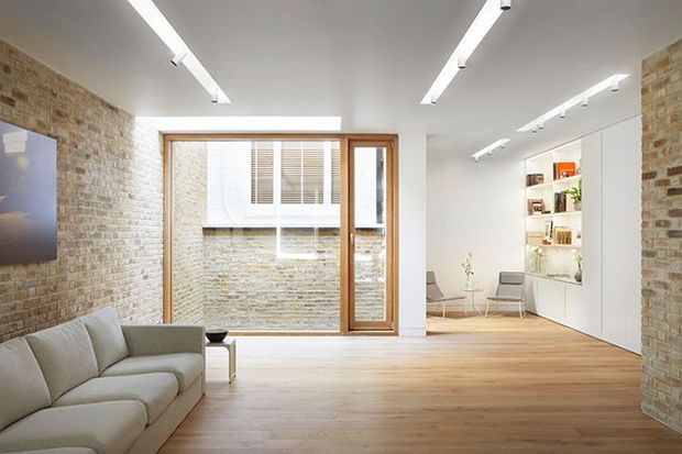 Bright contemporary open-plan living room with pale timber flooring, exposed light brick walls, a large timber-framed glazed opening to a brick courtyard, a light sectional sofa, and streamlined white built-in shelving and cabinetry. Linear ceiling-mounted lighting creates an airy gallery-like interior.