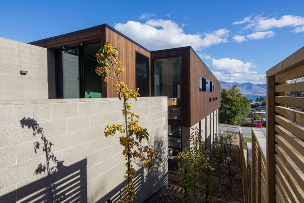 Exterior view of a contemporary multi-level residence on a sloped site, combining dark timber-clad upper volumes, large black-framed glazing, and pale concrete block walls. A timber privacy screen and planted landscaping frame the view, with a suburban street and mountain backdrop beyond.