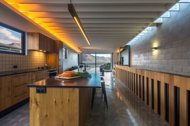 A long, narrow contemporary kitchen with a central timber island and dark benchtop, integrated cabinetry, tiled splashback, and exposed structural ceiling beams. A textured concrete-block wall with timber slatted detailing runs along the right side. Warm concealed lighting and linear pendant lighting create a welcoming atmosphere, while full-height glazing at the far end frames a mountainous landscape and water view.