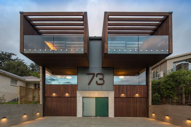 Street-facing contemporary residential facade with a strongly symmetrical composition. Two projecting upper volumes feature dark timber soffits, timber pergola-like roof screens, and glazed front sections. Below are paired timber-clad garage doors flanking a recessed central entry marked with the number 73. Low rendered masonry boundary walls, integrated exterior lighting, and neighbouring homes frame the driveway approach.