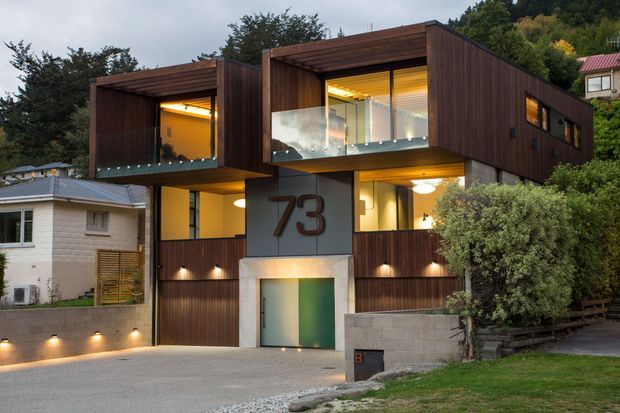 Contemporary multi-level residential house on a sloped suburban site, featuring dark timber-clad box forms, projecting glazed balconies, warm interior lighting, a recessed entry marked with the number 73, and an integrated driveway and retaining walls.