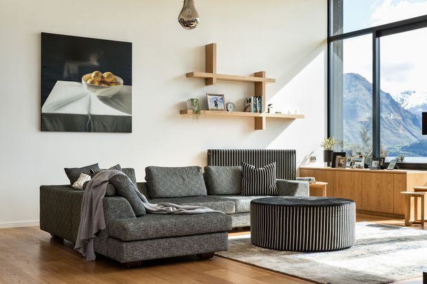 Bright contemporary living room with a large grey sectional sofa, striped round ottoman, timber flooring, a pale wall with sculptural floating timber shelving, and a low timber media/storage cabinet. Floor-to-ceiling black-framed glazing opens views toward a mountainous landscape, while a radiator and framed artwork add functional and decorative detail.