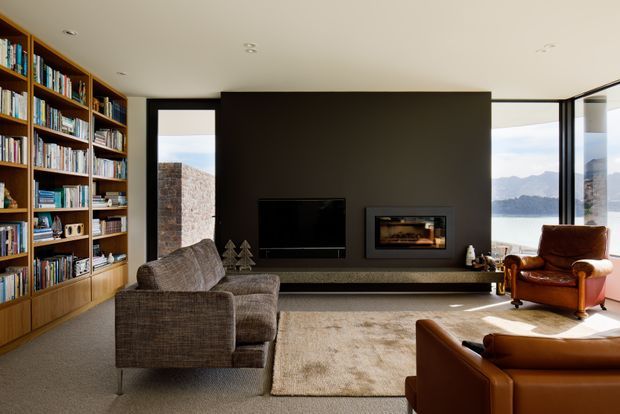A refined contemporary living room with a full-height built-in timber bookcase, large dark feature wall, flush-mounted television, and an inset fireplace. A textured grey sofa and brown leather lounge chairs sit on a large neutral rug. Floor-to-ceiling glazing at the right opens views toward a body of water and mountains, while exposed masonry is visible outside.