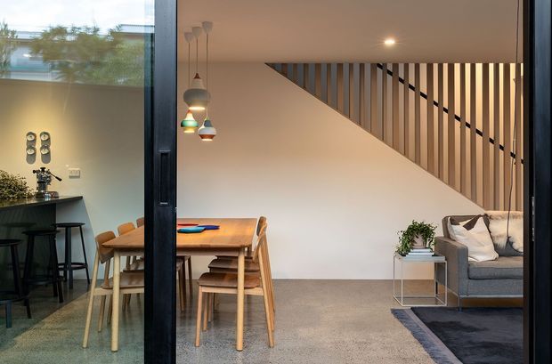 A contemporary open-plan interior viewed through a dark-framed glazed opening, with a compact kitchen and island on the left, a timber dining table in the centre, and a living area with a grey sofa on the right. A timber stair balustrade with vertical slats forms a strong architectural feature across the upper wall. The room has warm neutral finishes, carpeted flooring, timber furniture, and colourful pendant lights.