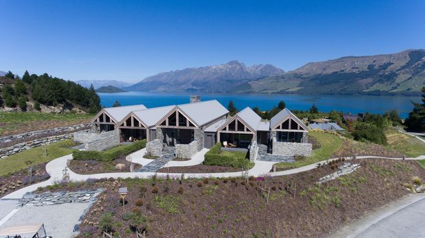 Aerial view of a low-rise residential lodge-style building set on a landscaped sloping site beside a deep blue lake, with rugged mountain ranges in the background. The building has multiple connected gabled volumes, pale metal pitched roofs, dark timber-framed glazing, and grey stone-clad walls. Curving paved walkways and terraced stone retaining walls surround the property.