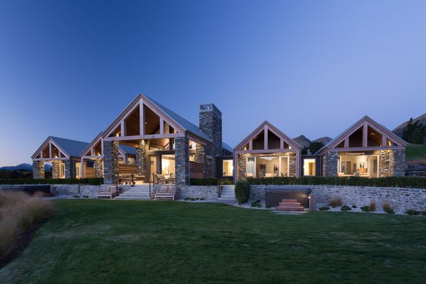 Exterior view of a contemporary alpine-style residential compound at dusk, composed of several connected or closely grouped gabled buildings. The façades combine extensive natural stone masonry with dark timber framing and large glazed openings. Warm interior lighting, landscaped lawns, low stone retaining walls, and a mountainous setting are visible.