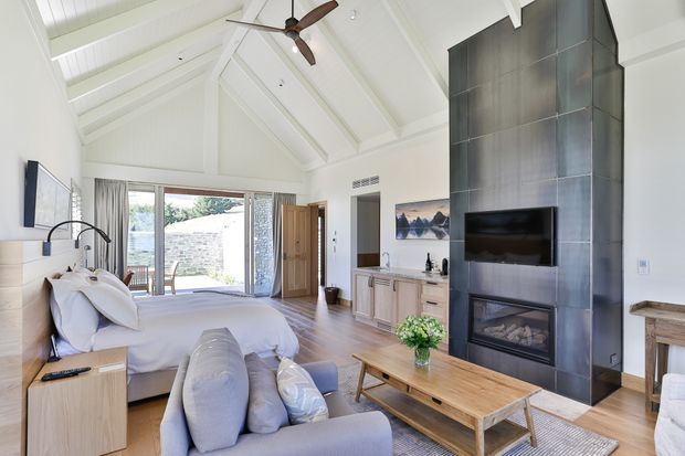 Bright open-plan bedroom and living area with a vaulted white timber-lined ceiling, exposed beams, ceiling fan, timber flooring, and large glazed doors opening to an outdoor courtyard. A bed occupies the left side, while a soft sofa, timber coffee table, and built-in dark tiled fireplace and TV feature wall define the living area.