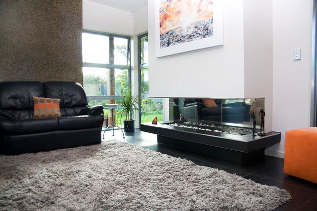 A bright contemporary living room with large corner windows, a black leather sofa, shag rug, orange accent ottoman and cushion, and a long mirrored fireplace feature with dark tiled flooring. Abstract artwork and indoor planting add colour and texture.