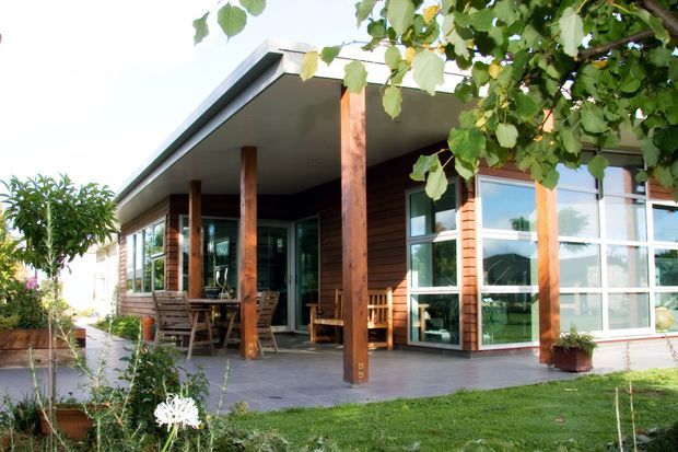 Single-storey contemporary house exterior with a broad flat projecting roof, deep covered veranda, warm horizontal timber cladding, large timber posts, and extensive multi-pane glazing. The building opens onto a paved outdoor seating area and landscaped garden, with timber outdoor furniture beneath the covered terrace.