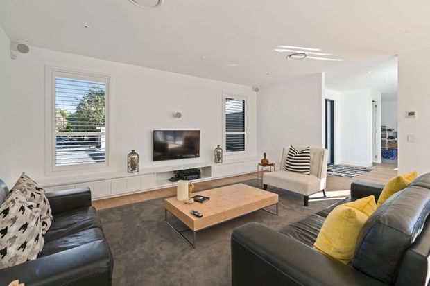 Bright contemporary living room with two black leather sofas, a neutral armchair, timber coffee table, wall-mounted television and extensive white built-in media cabinetry. White walls, plantation shutters, recessed lighting and large windows create an airy, clean interior.