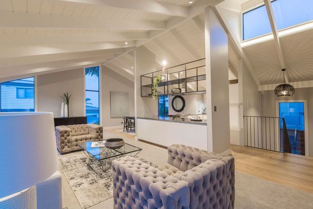 A bright, open-plan contemporary living area with a pair of tufted pale sofas, a glass coffee table, and a large patterned rug. The room features white painted timber-lined vaulted ceilings, exposed structural beams, tall windows, and skylights. An open black-framed partition separates the living space from a kitchen or dining zone, while a stair and mezzanine-like landing are visible to the right.