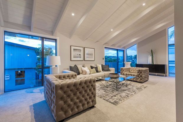 Bright, spacious living room with a dramatic white-painted exposed-beam vaulted ceiling, large windows and glazed doors, plush tufted seating, a neutral sofa, glass coffee table, patterned area rug, and framed artwork. The room has a refined contemporary feel and overlooks a blue-toned exterior setting.