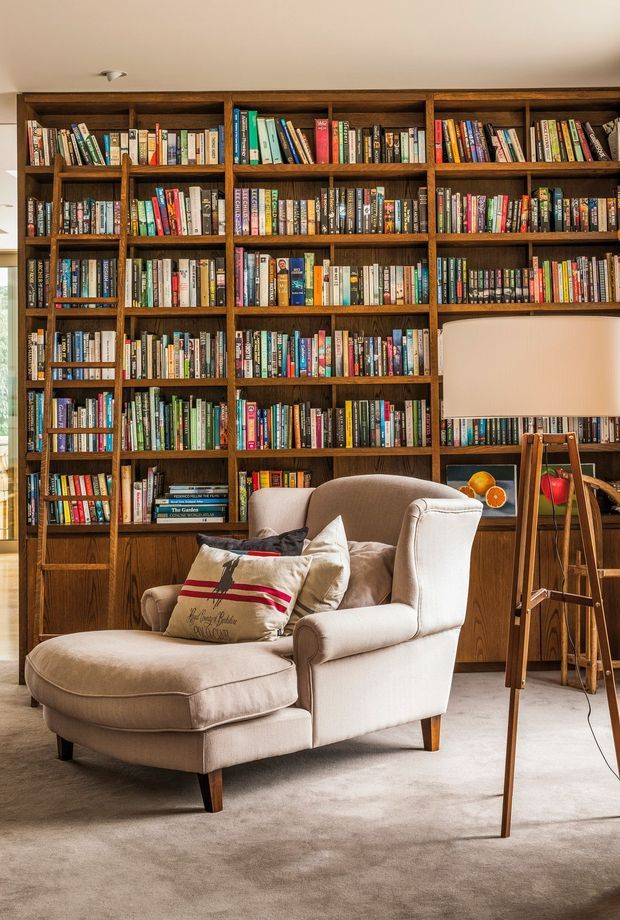 A warm, traditional reading room with a wall-to-wall dark timber built-in bookcase filled with books. In front is a plush cream upholstered armchair with matching ottoman and cushions, alongside a timber tripod floor lamp and small framed artwork. Soft natural light, beige carpet, and warm wood tones create a cozy library atmosphere.