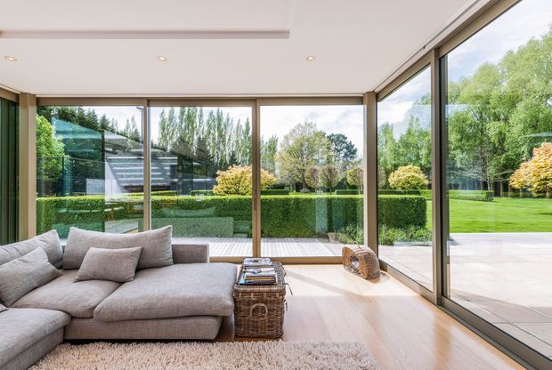 Bright contemporary living room with a large neutral sectional sofa, low woven coffee table, pale timber flooring, recessed ceiling lights, and expansive corner glazing overlooking a manicured garden and patio. The room has a calm, airy indoor-outdoor connection with slim dark window frames and minimal detailing.