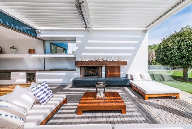 Bright modern indoor-outdoor living room with a white louvred pergola ceiling, full-height glazing, built-in white cabinetry and shelving, a central timber-and-stone fireplace, and a furnished timber deck extending toward a manicured garden. The space features low-profile white sofas, a striped accent cushion, a timber coffee table, and strong linear shadows from the overhead structure.